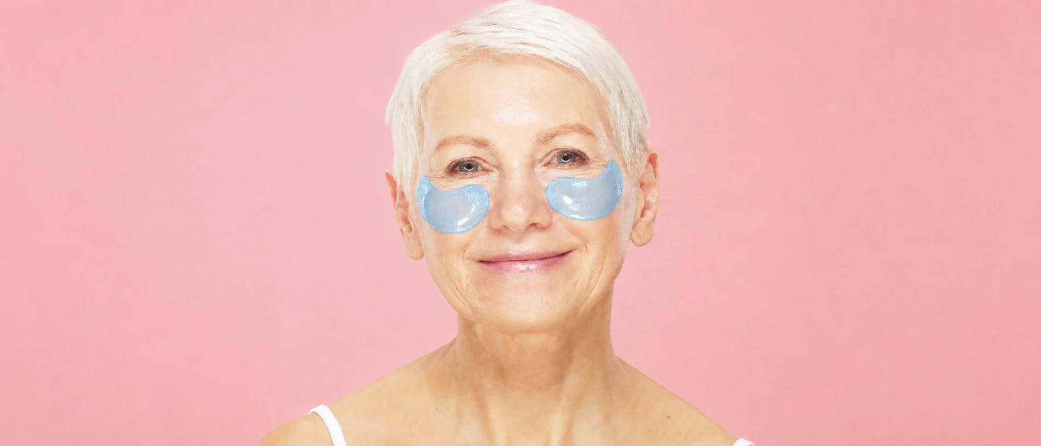 Smiling mature woman wearing blue hydrogel under-eye patches against a pink background, a needle-free way to care for under-eye fine lines.