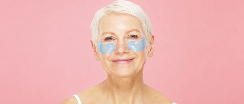 Smiling mature woman wearing blue hydrogel under-eye patches against a pink background, a needle-free way to care for under-eye fine lines.