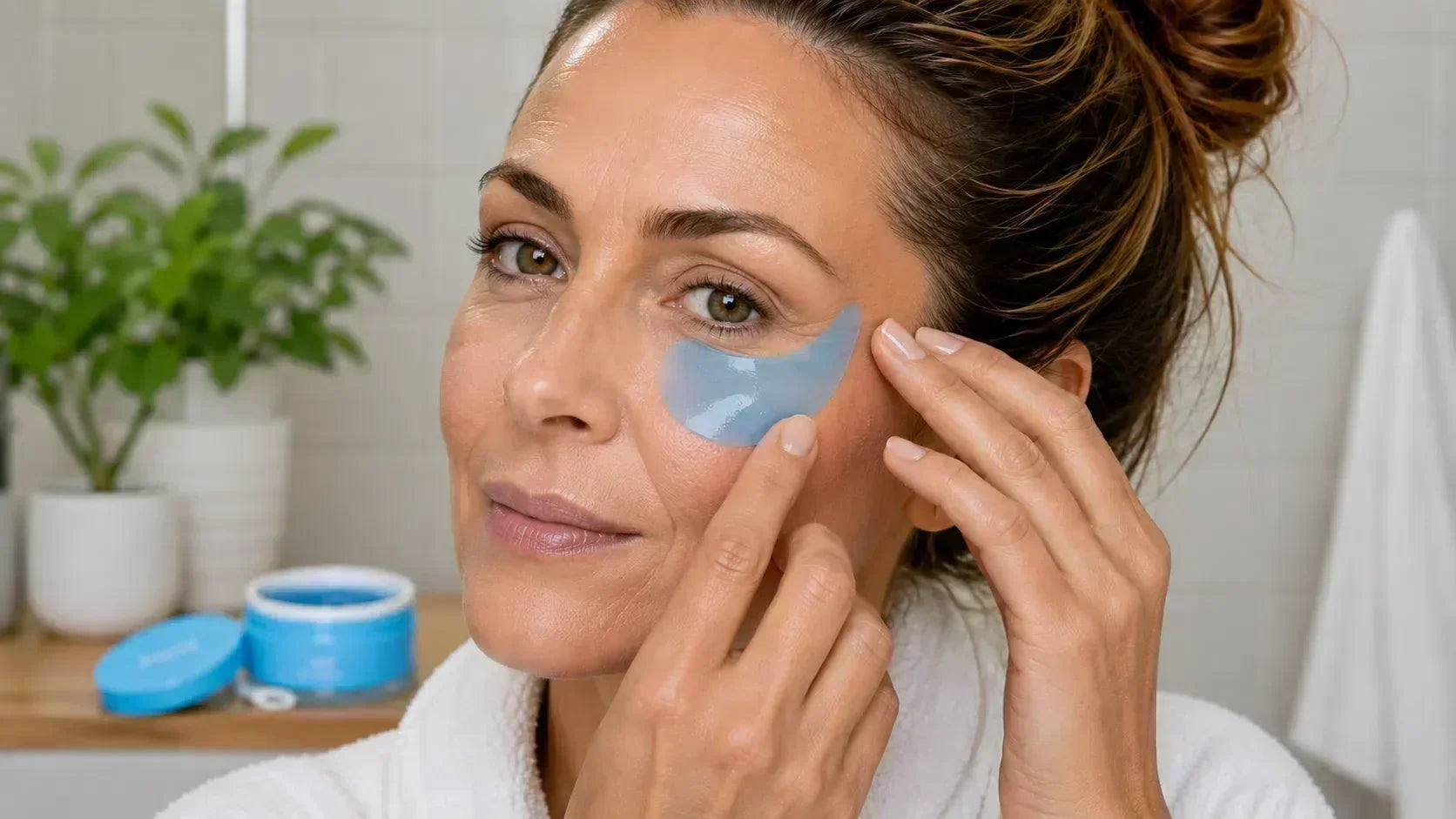 Woman over 40 applying an Aloha Blue hydrogel under-eye patch in a bright bathroom, Opuola under-eye care