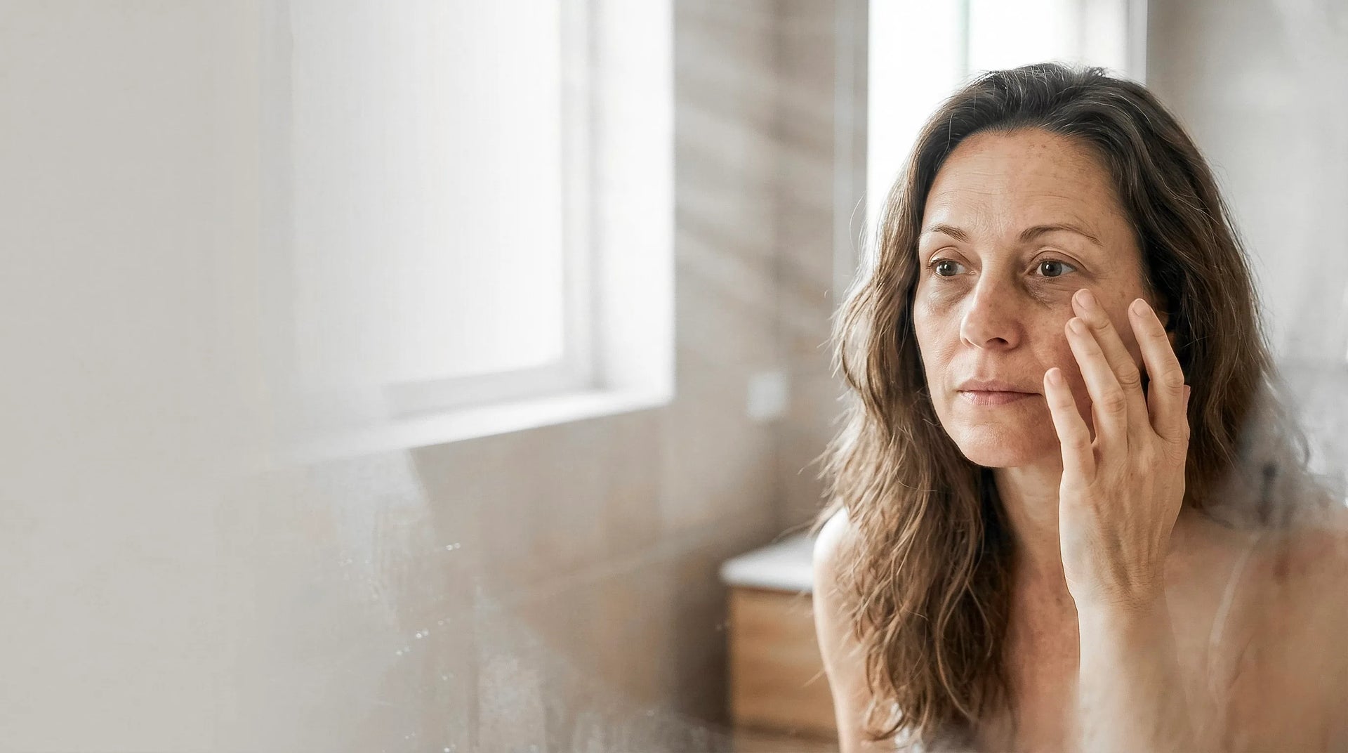 Woman in her mid-40s examining the skin under her eyes in a bathroom mirror during morning light