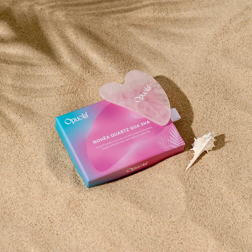 Opuola Nohéa rose quartz gua sha facial sculpting tool displayed on sand with tropical inspired packaging
